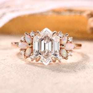 May include: A rose gold ring featuring a large, faceted, hexagonal-cut clear gemstone as the centerpiece. The ring is accented with small opalescent gemstones and clear accent stones, set around the main stone. The ring has a delicate, elegant design.