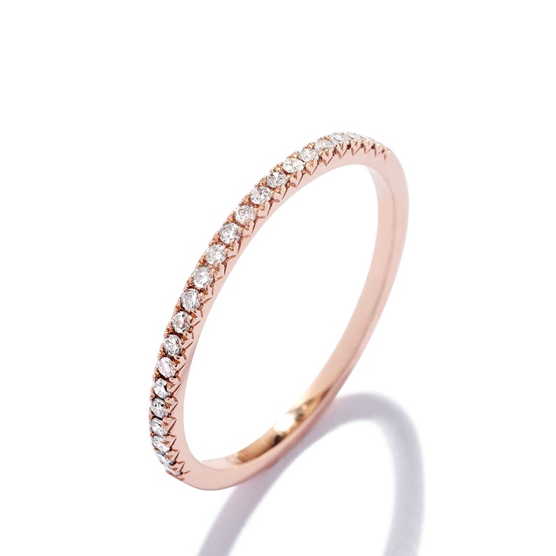 May include: A thin rose gold band ring featuring a continuous row of small, round, clear pav&eacute;-set diamonds along the top half of the band. The metal has a polished finish, and the stones are set in a shared prong setting style.