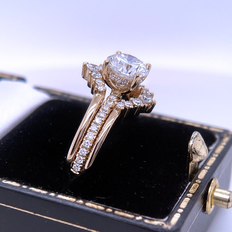 May include: A gold engagement ring set with a large, round, clear gemstone and smaller accent stones. The set includes a curved band and a matching ring, all displayed in a black ring box.