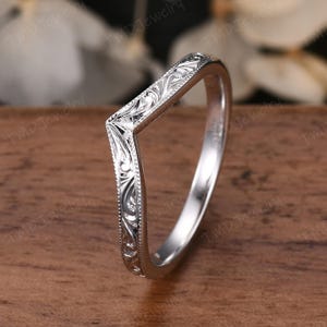 May include: A silver-toned V-shaped ring featuring intricate engraved scrollwork patterns along the outer band. The metal surface has a polished finish with milgrain detailing along the edges, creating a distinct chevron silhouette for the band.