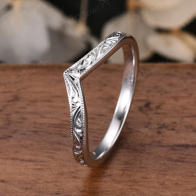 May include: A silver-toned V-shaped ring featuring intricate engraved scrollwork patterns along the outer band. The metal surface has a polished finish with milgrain detailing along the edges, creating a distinct chevron silhouette for the band.