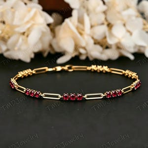 May include: A gold-colored bracelet with a series of rectangular links and small, round, red gemstones. The bracelet is displayed on a dark surface with white flowers in the background. The text "TMD Jewelry" is visible.