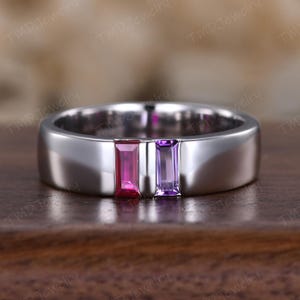 May include: A silver ring with a flat band design, featuring a rectangular pink gemstone and a rectangular purple gemstone set side by side. The ring is displayed on a wooden surface.