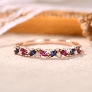 May include: Rose gold ring with a band of alternating marquise-cut gemstones. The gemstones include blue sapphires, rubies, white opals, and small diamonds. The ring is set on a neutral background.