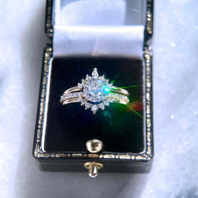 May include: A gold engagement ring set with a large, round, clear gemstone, surrounded by smaller stones in a floral design. The ring sits in a black jewelry box with a gold clasp, against a soft, white background.