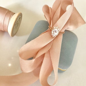 May include: A delicate engagement ring with a large oval-shaped center stone surrounded by smaller diamonds, set in a gold band. The ring is displayed in a light blue velvet ring box with a blush pink satin ribbon tied in a bow.