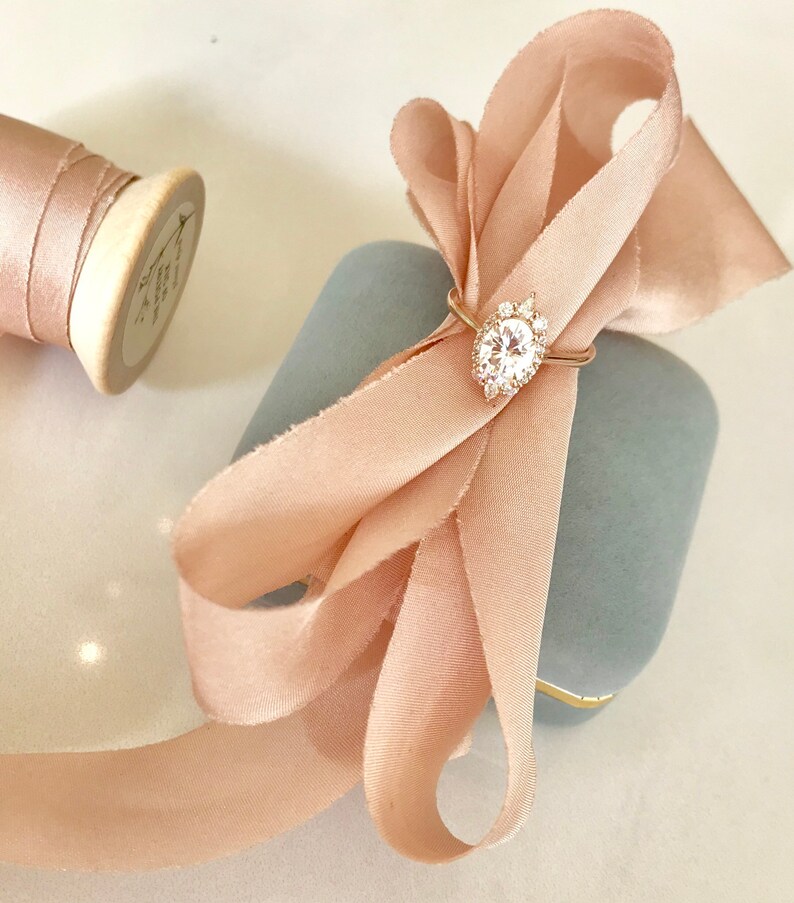 May include: A delicate engagement ring with a large oval-shaped center stone surrounded by smaller diamonds, set in a gold band. The ring is displayed in a light blue velvet ring box with a blush pink satin ribbon tied in a bow.