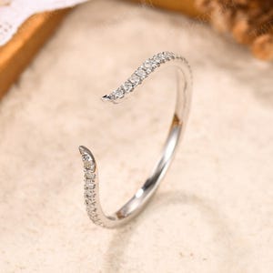 May include: A silver-toned open ring featuring a delicate band adorned with a row of small, clear, sparkling stones along the top and bottom curved ends. The metal has a polished, reflective finish, creating a bright and clean appearance.