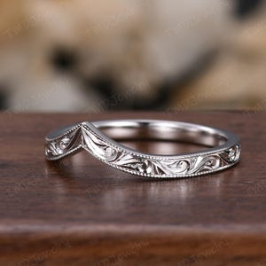 May include: A silver-toned chevron-shaped ring featuring intricate engraved scrollwork patterns along the band. The metal surface displays a polished finish with detailed filigree carvings and a beaded milgrain edge bordering the design.