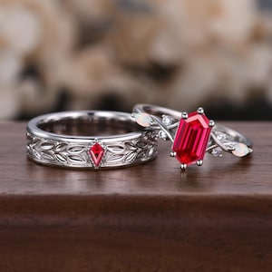 May include: A pair of silver wedding bands with intricate designs. The engagement ring features a large, red, octagon-shaped gemstone and smaller white gemstones. The wedding band has a red gemstone in the center.