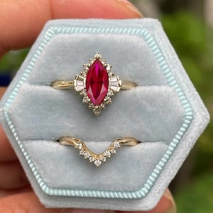 May include: A marquise-cut ruby engagement ring with a halo of small diamonds and baguette-cut stones. A matching curved band with small diamonds is below. Both rings are gold and set in a light blue velvet box.