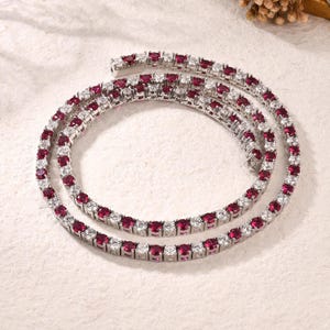 May include: A silver-toned necklace with alternating red and clear gemstones. The necklace is composed of multiple strands, each featuring a continuous line of round-cut stones. The gemstones are set closely together, creating a sparkling, elegant appearance.