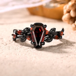 May include: A black ring with a coffin-shaped, deep red gemstone as the centerpiece. The ring features a floral design with smaller red gemstones and black leaves. The ring is set against a neutral background, highlighting its gothic aesthetic.