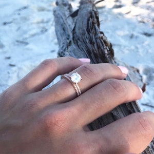May include: A close-up of a rose gold engagement ring with a round, clear center stone and a band of smaller stones. The ring is worn on the finger of a person's hand.