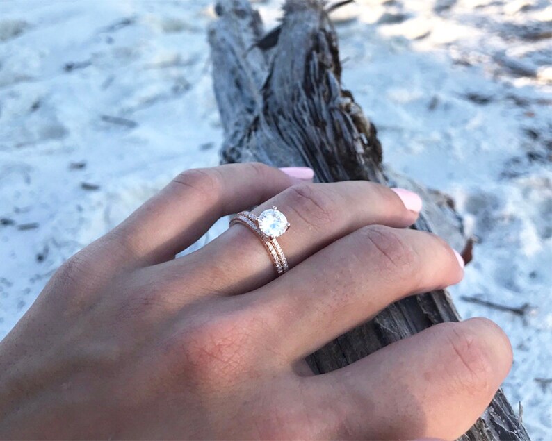 May include: A close-up of a rose gold engagement ring with a round, clear center stone and a band of smaller stones. The ring is worn on the finger of a person's hand.