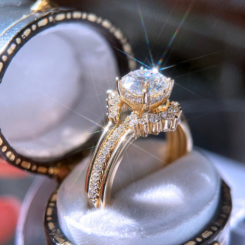 May include: A gold engagement ring with a large, clear, round-cut diamond as the centerpiece. The ring features a halo of smaller diamonds around the main stone and along the band, set in a gold band. The ring is displayed in a velvet-lined ring box.