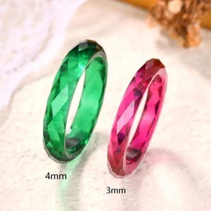 May include: Two faceted rings, one emerald green and one vibrant pink, are displayed side by side. The green ring is labeled with a 4mm width, and the pink ring is labeled with a 3mm width. Both bands feature a geometric, multi-faceted surface.