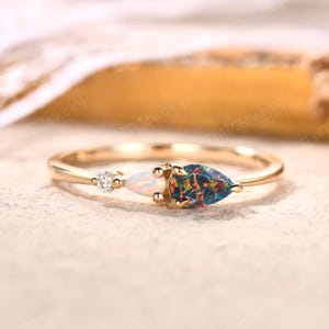 May include: A gold ring featuring a pear-shaped dark opal with iridescent flashes, a smaller pear-shaped white opal, and a single round brilliant-cut diamond. The stones are set in a delicate gold band with a polished finish and prong settings.