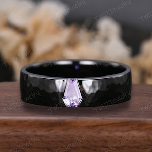 Puede incluir: Un anillo negro con textura martillada y una piedra preciosa de color púrpura claro. El anillo tiene una banda ancha y una piedra preciosa de forma geométrica. El anillo se muestra sobre una superficie de madera. La marca "TMD Jewelry" es visible.