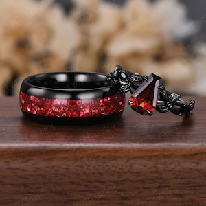 May include: Two black rings. One ring features a band filled with red crushed stone. The other ring has a black floral design and a large, rectangular, red gemstone. The rings are displayed on a wooden surface.