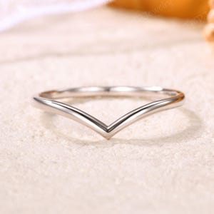 May include: A silver-colored ring with a V-shaped design. The ring is made of a shiny, reflective metal and sits on a light-colored surface. The ring's simple, elegant design makes it suitable for various occasions.