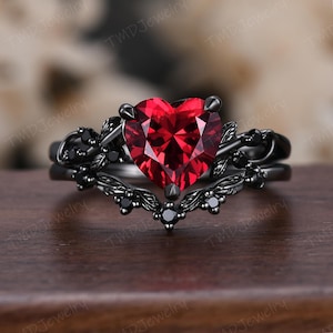 Puede incluir: Un conjunto de anillos de compromiso negros con una piedra roja facetada en forma de corazón. El anillo está adornado con piedras negras y detalles florales intrincados. El conjunto incluye una banda a juego.