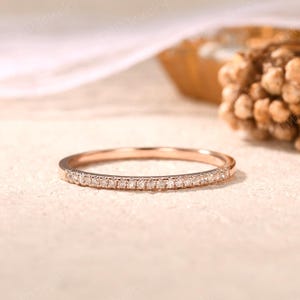 May include: A thin rose gold band ring featuring a row of small, clear, round-cut stones set along the front half of the band. The metal has a polished finish, and the stones are arranged in a continuous line, creating a delicate, shimmering appearance.