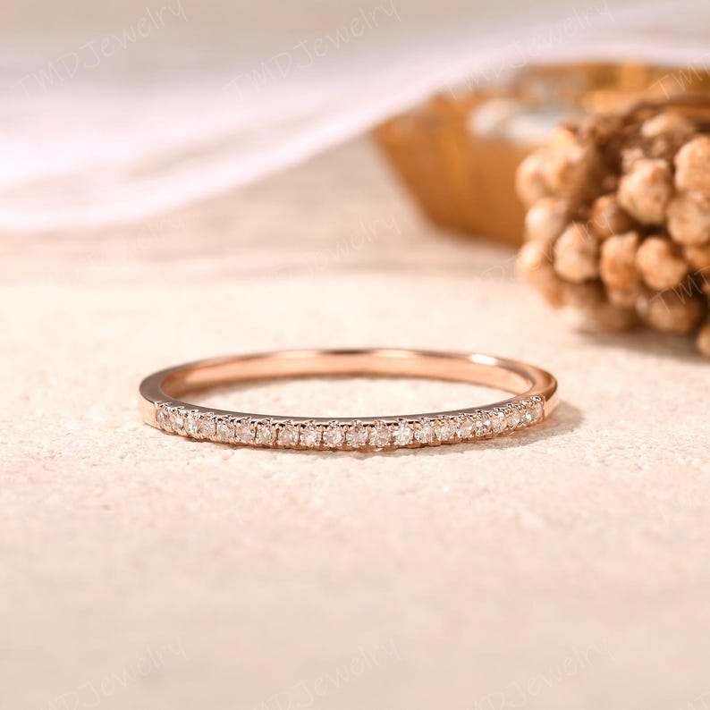 May include: A thin rose gold band ring featuring a row of small, clear, round-cut stones set along the front half of the band. The metal has a polished finish, and the stones are arranged in a continuous line, creating a delicate, shimmering appearance.