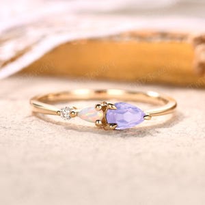 May include: A gold ring featuring three gemstones: a small diamond, an opal, and a pear-shaped lavender gemstone. The ring has a delicate band and is set on a neutral surface.