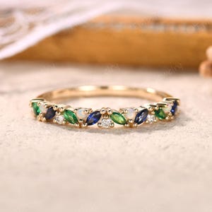 Może przedstawiać: Złoty pierścionek z rzędem naprzemiennych kamieni szlachetnych o szlifie markizy. Kamienie szlachetne obejmują zielone, niebieskie i białe kamienie, tworząc kolorowy i elegancki wzór. Pierścionek leży na neutralnej powierzchni.