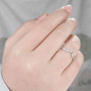 May include: A silver-toned chevron-shaped ring featuring an intricate engraved scrollwork pattern along the band. The ring has a pointed V-shape design and a polished metallic finish, worn on the ring finger of a hand.