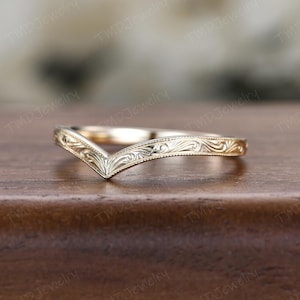 May include: A gold V-shaped ring featuring an intricate engraved scrollwork pattern along the band. The metal surface displays a polished finish with detailed filigree-style carvings that follow the chevron contour of the jewelry piece.