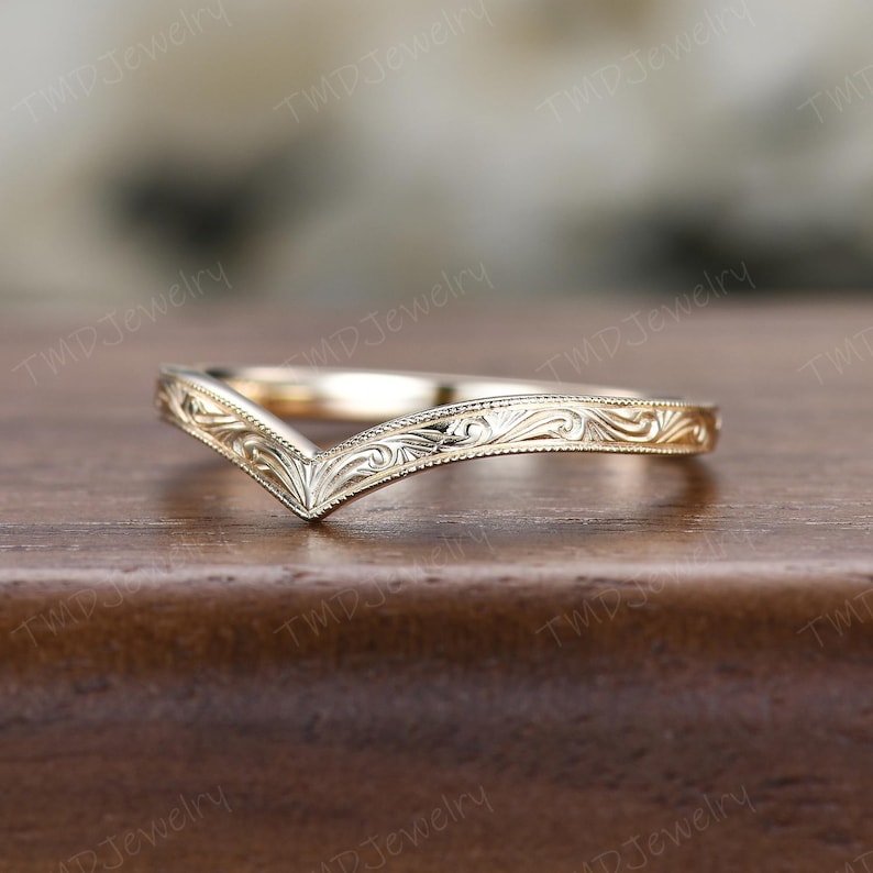 May include: A gold V-shaped ring featuring an intricate engraved scrollwork pattern along the band. The metal surface displays a polished finish with detailed filigree-style carvings that follow the chevron contour of the jewelry piece.