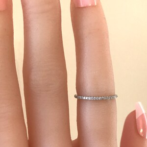 May include: A thin, silver-toned metal band ring featuring a continuous row of small, clear, sparkling stones set along the entire circumference. The ring is worn on a finger, showcasing its delicate, minimalist design and reflective metallic finish.