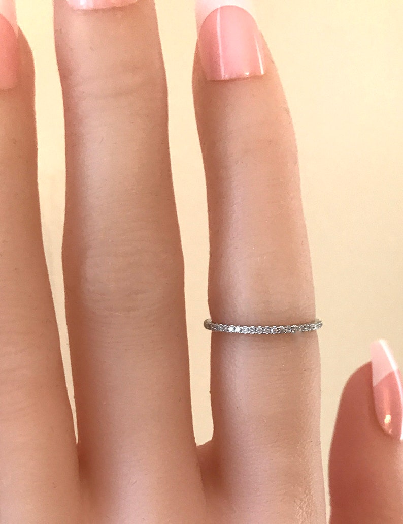 May include: A thin, silver-toned metal band ring featuring a continuous row of small, clear, sparkling stones set along the entire circumference. The ring is worn on a finger, showcasing its delicate, minimalist design and reflective metallic finish.