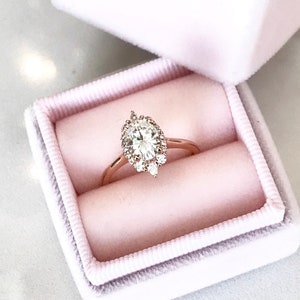 May include: A rose gold engagement ring with a large oval-shaped diamond surrounded by a halo of smaller diamonds. The ring is in a pink velvet ring box.