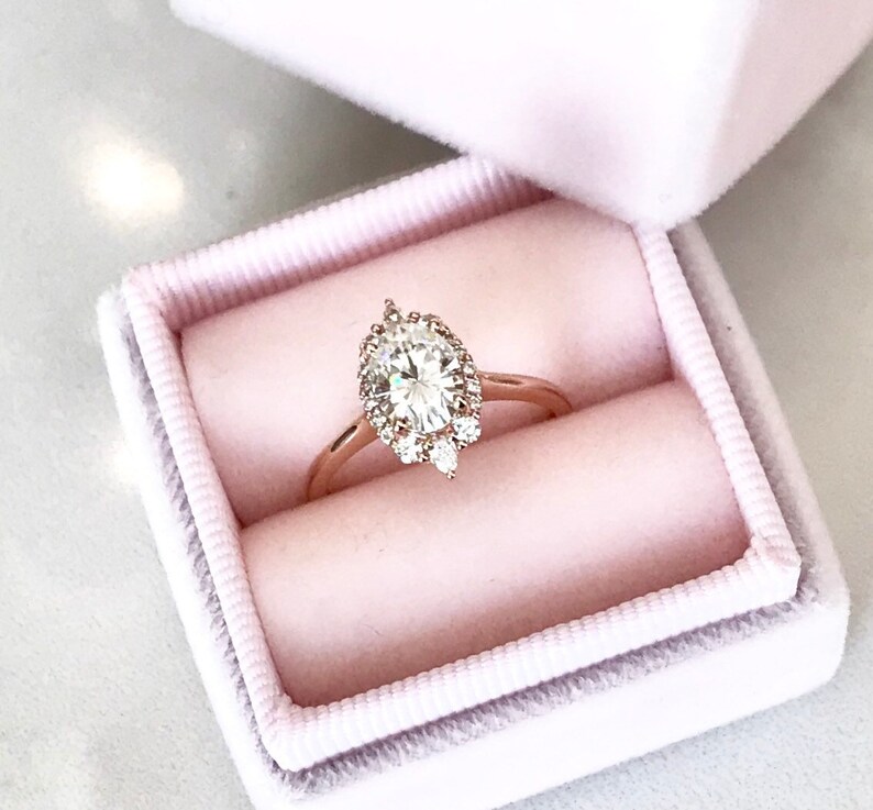 May include: A rose gold engagement ring with a large oval-shaped diamond surrounded by a halo of smaller diamonds. The ring is in a pink velvet ring box.