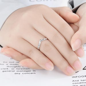May include: A silver-toned chevron-shaped ring featuring an intricate engraved floral pattern along the band. The ring has a pointed V-shape design and a polished metallic finish, worn on the ring finger of a hand resting on a printed surface.