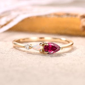 May include: A delicate gold ring featuring three gemstones. A small diamond, an opal, and a pear-shaped ruby are set in a row. The ring has a thin band and is displayed on a neutral surface.