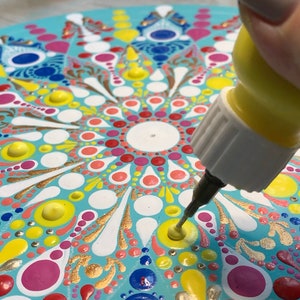 May include: A hand holding a yellow bottle of glue with a white tip, squeezing glue onto a colorful mandala painted on a round wooden surface.