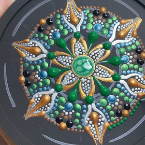May include: A close-up of a round, black coaster with a green, gold, and white mandala design. The mandala is made of dots and lines, and there is a green gem in the center. A bottle of purple glue is being used to add more dots to the design.