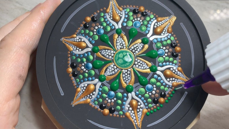 May include: A close-up of a round, black coaster with a green, gold, and white mandala design. The mandala is made of dots and lines, and there is a green gem in the center. A bottle of purple glue is being used to add more dots to the design.