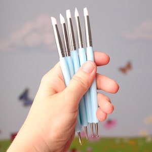 May include: A hand holding a set of six light blue and silver double-ended dotting tools. Each tool has a white tip on one end and a metal tip on the other. The background is a soft, blurred scene with butterflies.