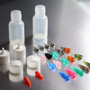 May include: Two clear plastic bottles with white caps and a variety of colored and clear plastic needle tips. The needle tips are arranged in rows on a silver surface.