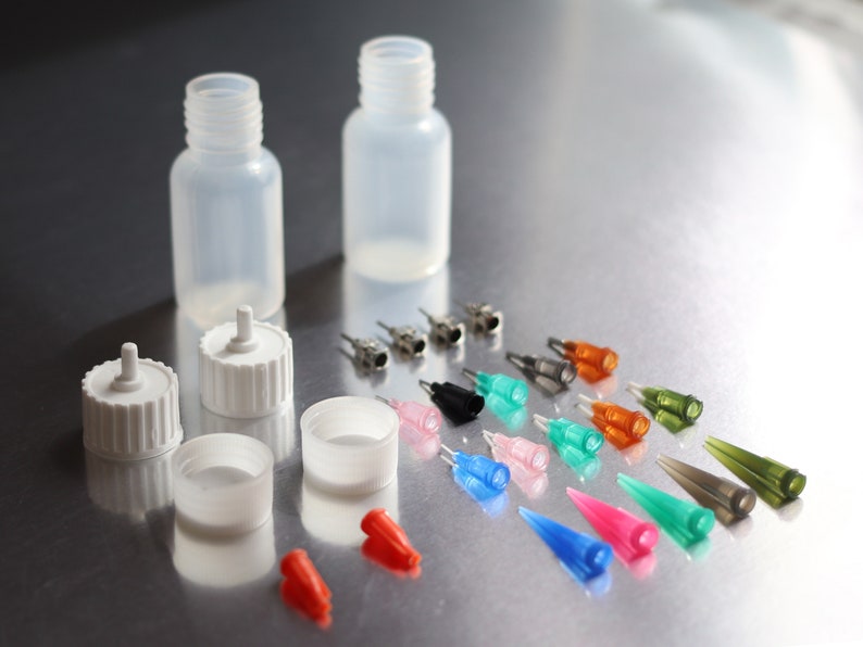May include: Two clear plastic bottles with white caps and a variety of colored and clear plastic needle tips. The needle tips are arranged in rows on a silver surface.