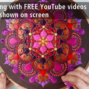 Puede incluir: Primer plano de una tabla redonda de madera con un dise&ntilde;o de mandala colorido pintado sobre ella. El mandala presenta una variedad de colores, incluyendo rosa, rojo, naranja, dorado y morado. El dise&ntilde;o es intrincado y detallado, con una variedad de formas y patrones. La tabla est&aacute; rodeada por una superficie blanca con una paleta de colores de pintura y un conjunto de herramientas para pintar con puntos. El texto "TOOL 12" es visible en la esquina superior derecha de la imagen.