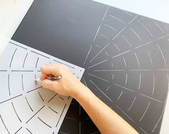 Puede incluir: Una persona utiliza una plantilla para trazar un diseño de telaraña en un lienzo negro. La plantilla es de color gris claro y el lienzo es de color gris oscuro.