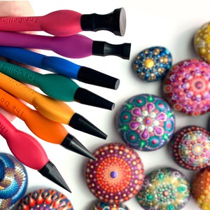 Puede incluir: Un conjunto de seis herramientas de punteado de colores con puntas negras, etiquetadas como "The Dotting Co.", que se utilizan para crear arte de puntos en rocas. Las herramientas est&aacute;n dispuestas en una mano, con las puntas hacia afuera. El fondo muestra varias rocas pintadas con dise&ntilde;os de mandalas coloridos.
