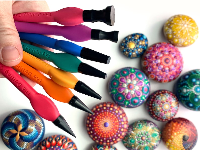 Puede incluir: Un conjunto de seis herramientas de punteado de colores con puntas negras, etiquetadas como "The Dotting Co.", que se utilizan para crear arte de puntos en rocas. Las herramientas est&aacute;n dispuestas en una mano, con las puntas hacia afuera. El fondo muestra varias rocas pintadas con dise&ntilde;os de mandalas coloridos.