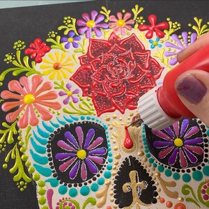 May include: A close-up of a sugar skull painting in progress. The skull is decorated with colorful flowers and dots. A hand is holding a bottle of red paint, adding details to the skull.